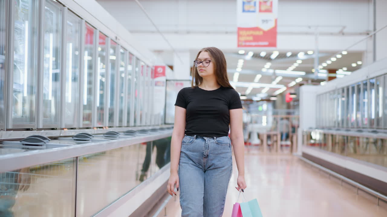 Kvinde, der shopper i sort top, går forbi et udstillingsvindue med en anden kunde i baggrunden. Hun holder indkøbsposer og kigger fremad, mens hun bevæger sig gennem et lyst og moderne butikscenter.