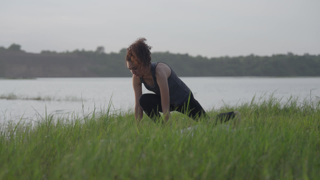 A slow-motion of a South Asian Woman in a low lunge yoga pose (Anjaneyasana) looking up, by a lake in tall grass