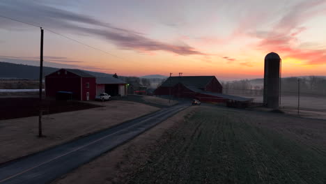 Truck-drives-on-rural-road-past-farm-buildings-and-barn-in-morning-sunrise