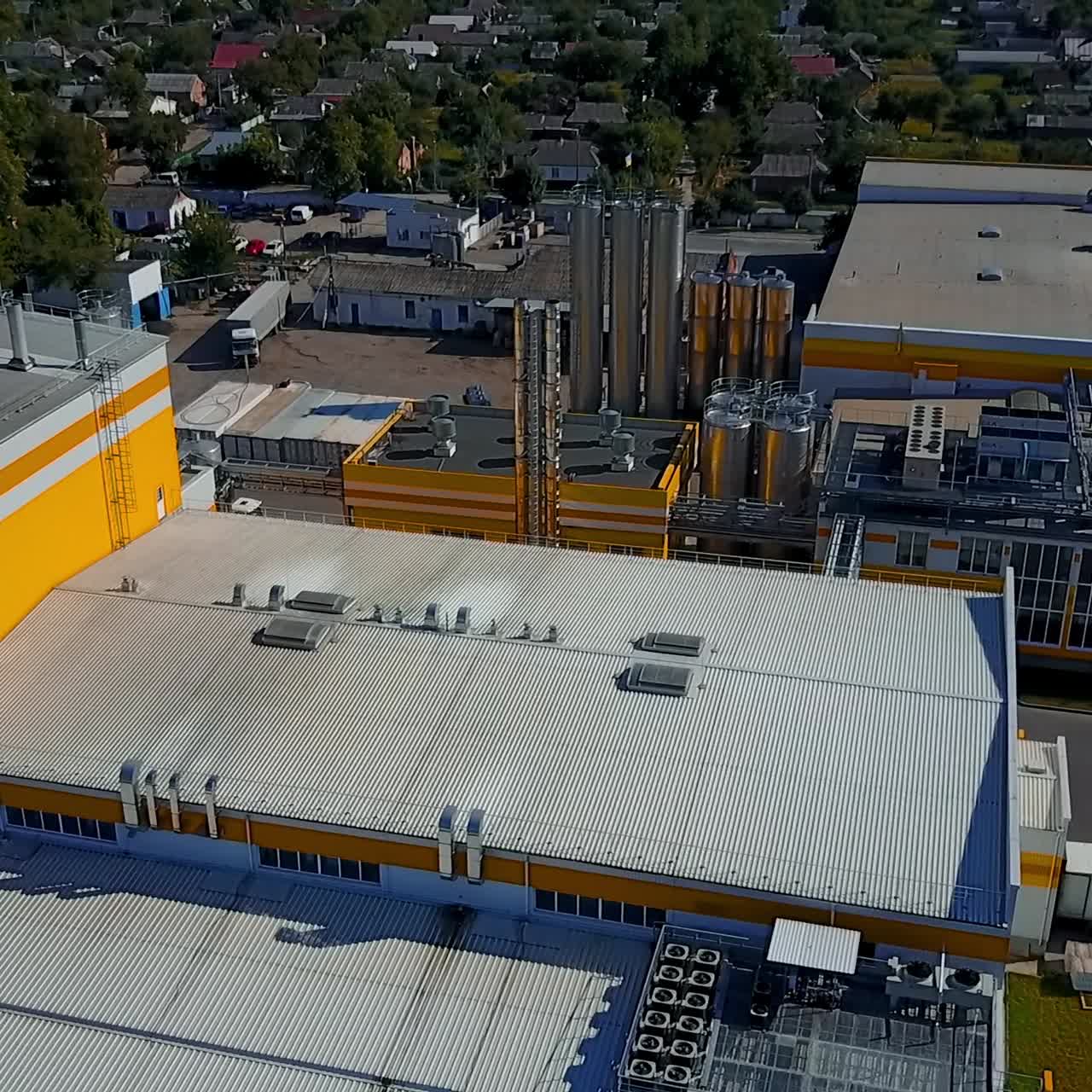Territory of the plant with silver pipes. Private houses and green trees at backdrop. Aerial view
