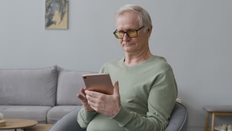 Lächelnder-älterer-Mann,-Der-Ein-Tablet-Verwendet,-Während-Er-Zu-Hause-In-Einem-Modernen-Wohnzimmer-Sitzt-1
