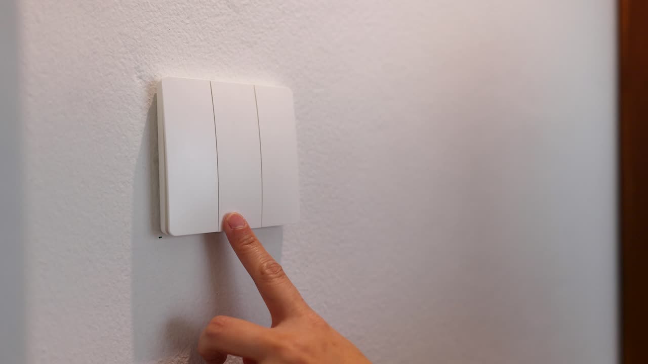 A close-up shot shows a finger pressing a white light switch on a plain wall