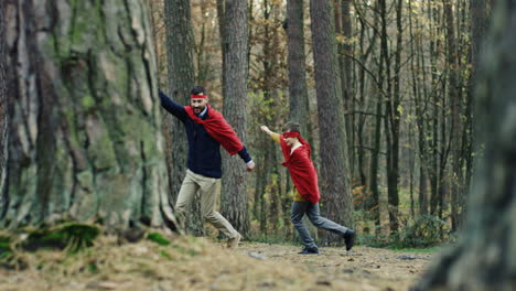 padre e hijo caucásicos alegres con capas rojas corriendo en el bosque y jugando a ser superhéroes