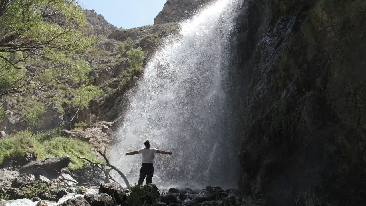 Man Stands Before a Powerful Waterfall with Arms Outstretched