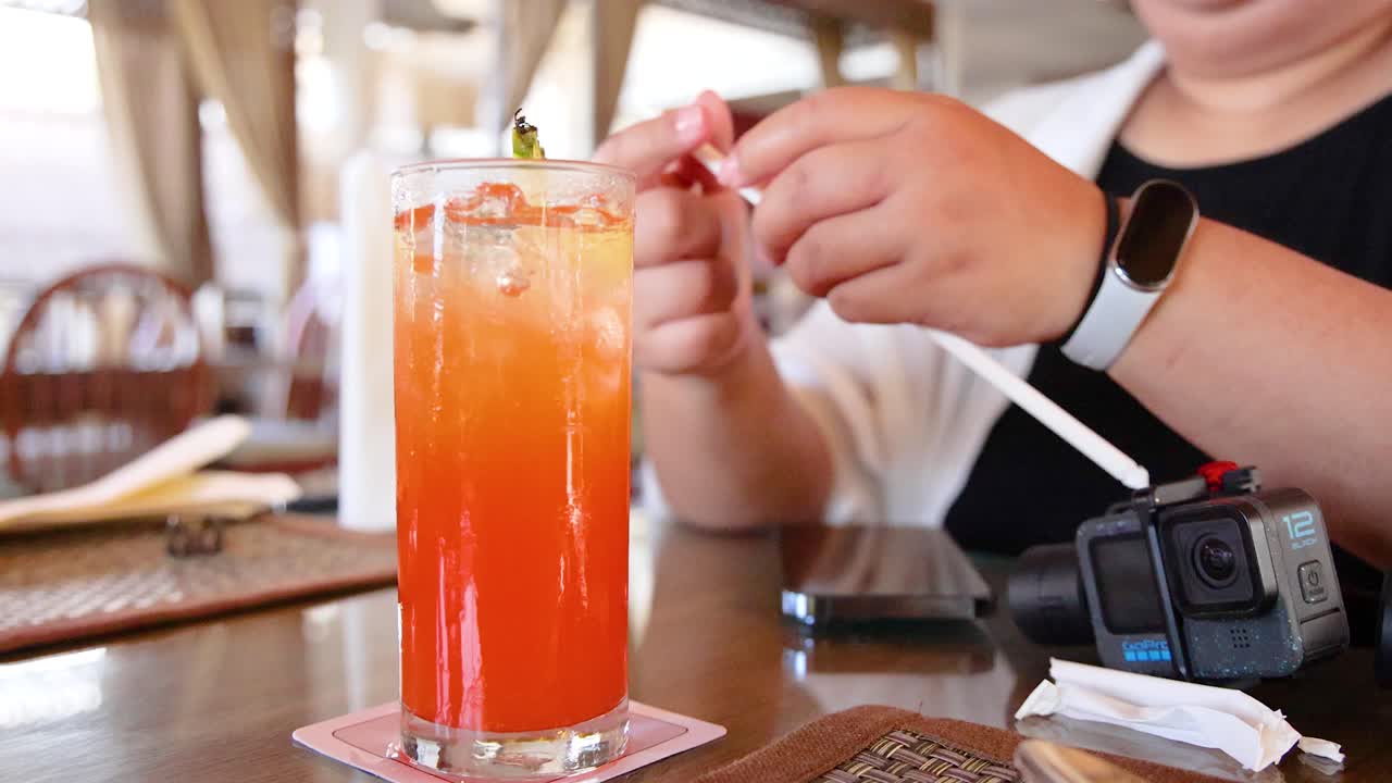 A person prepares a colorful fruity cocktail at a table, using a straw, in a bright, relaxed setting