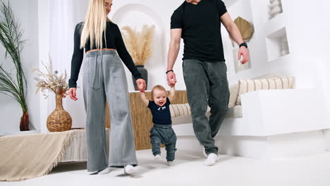 couple holding a baby by hands walk by the room. cute happy baby learning to walk, making first steps. low angle view.
