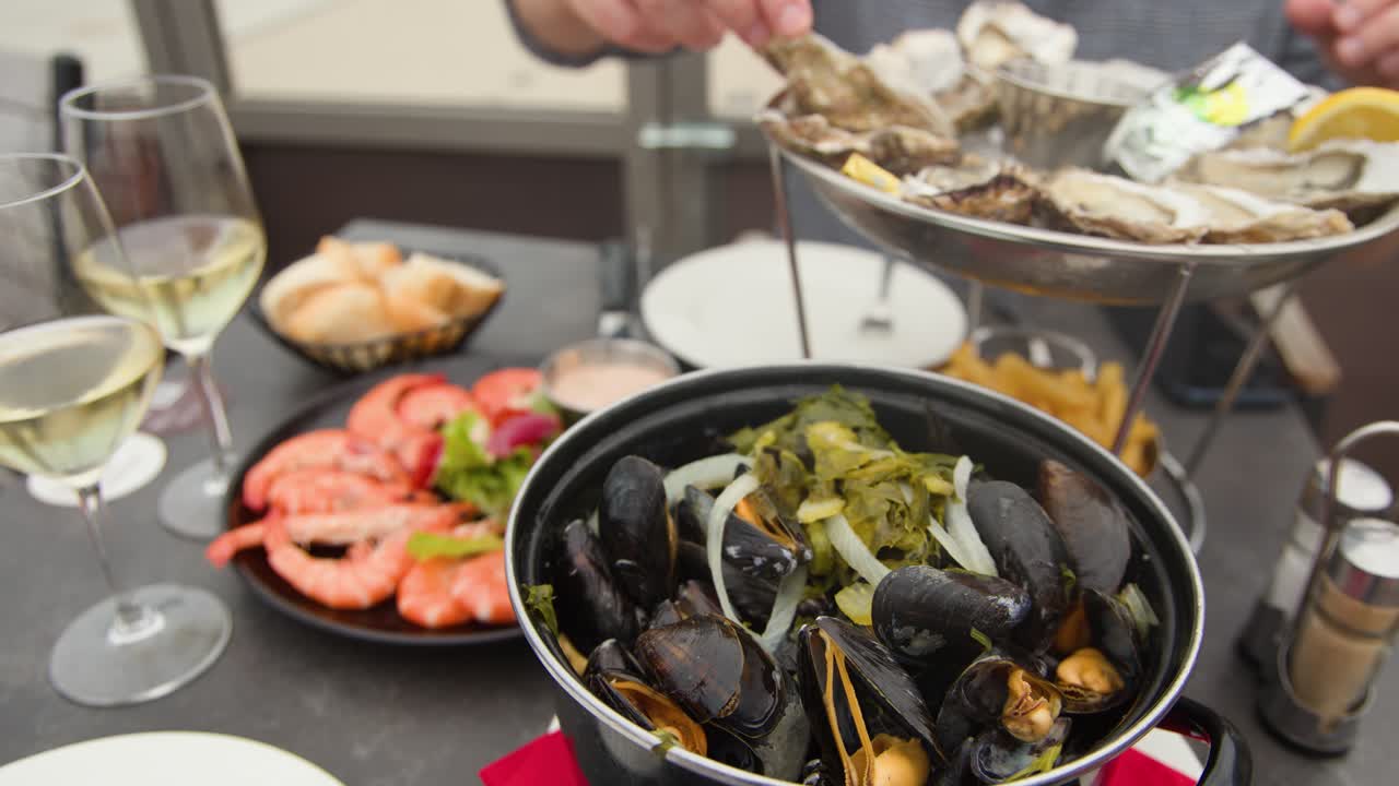 Person serves oysters and mussels at sunlit coastal restaurant table with shrimp, wine, and seafood