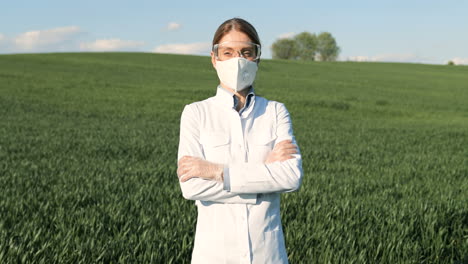 Mujer-Investigadora-Caucásica-Con-Bata-Blanca-Y-Gafas-Mirando-La-Cámara-Con-Los-Brazos-Cruzados-En-El-Campo-Verde