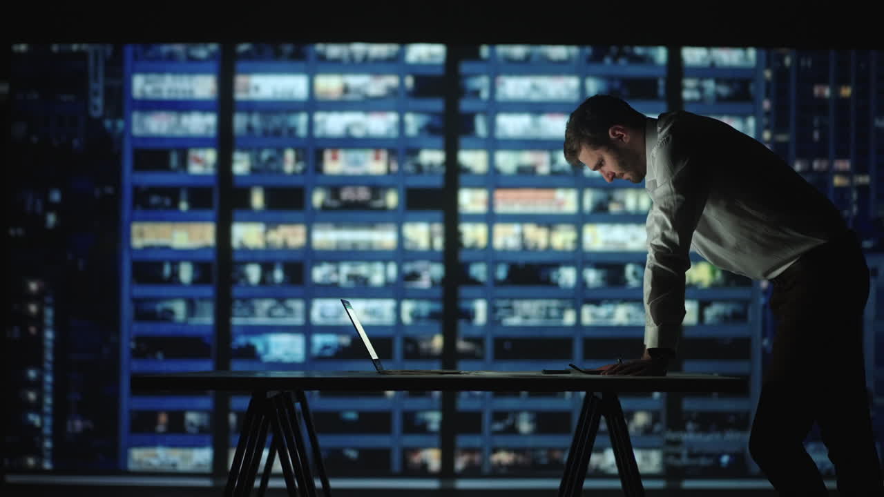 Tired young man working on a laptop late night in the office. Sleepy Businessman sitting at desk in dark office. Tired and stressed businessman in glasses works on a laptop of the night city office