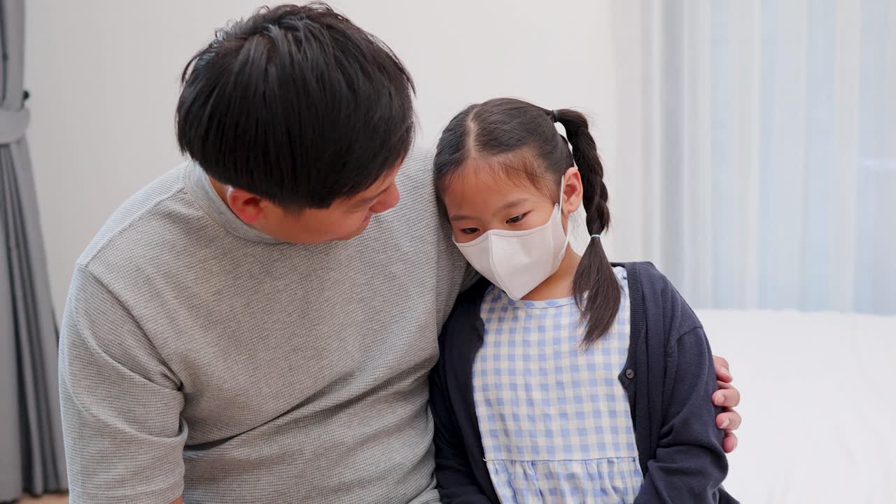 Father gently comforts ill daughter in mask, sitting together on bed in softly lit room