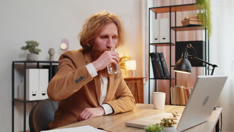 business man sitting at workplace desk drinking water while working with laptop in home office