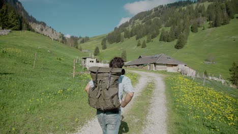 handheld, back of backpacker young man hiking on rural mountain valley dirt road