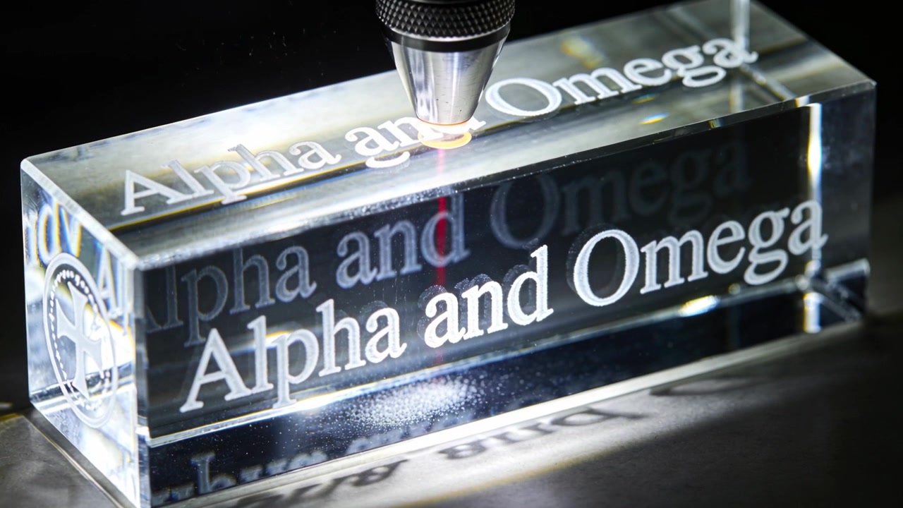 La testa laser si attiva, incidendo un blocco trasparente per definire il testo Alfa e Omega su un banco da lavoro