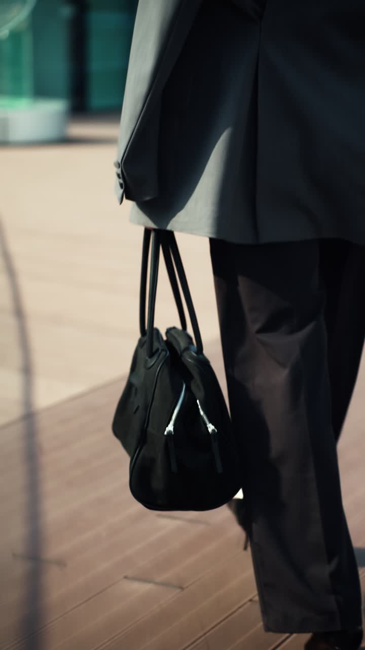 Close-up of a businesswoman walking in slow motion on a modern office terrace, carrying a black handbag - rear view