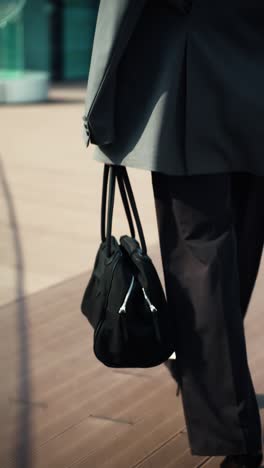 close-up of a businesswoman walking in slow motion on a modern office terrace, carrying a black handbag - rear view