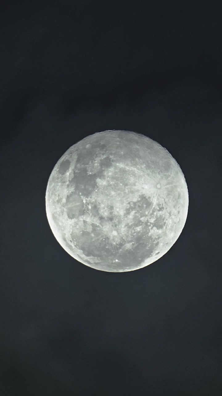 Close-up vertical de uma Lua Cheia brilhante contra um céu noturno escuro, revelando crateras lunares detalhadas enquanto nuvens finas passam pela lua