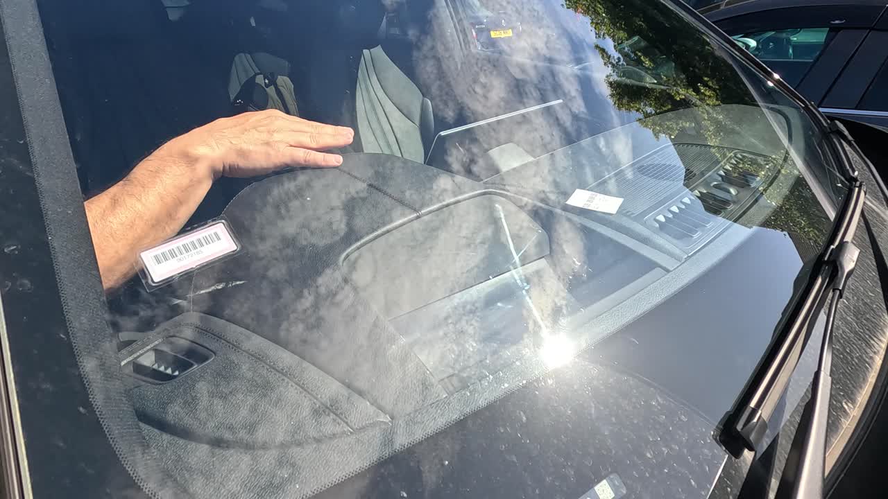 A man’s arm reaches through a car window to place a parking ticket on the dashboard in bright daylight, viewed from outside the vehicle