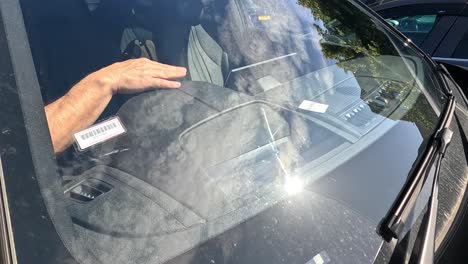 a man’s arm reaches through a car window to place a parking ticket on the dashboard in bright daylight, viewed from outside the vehicle