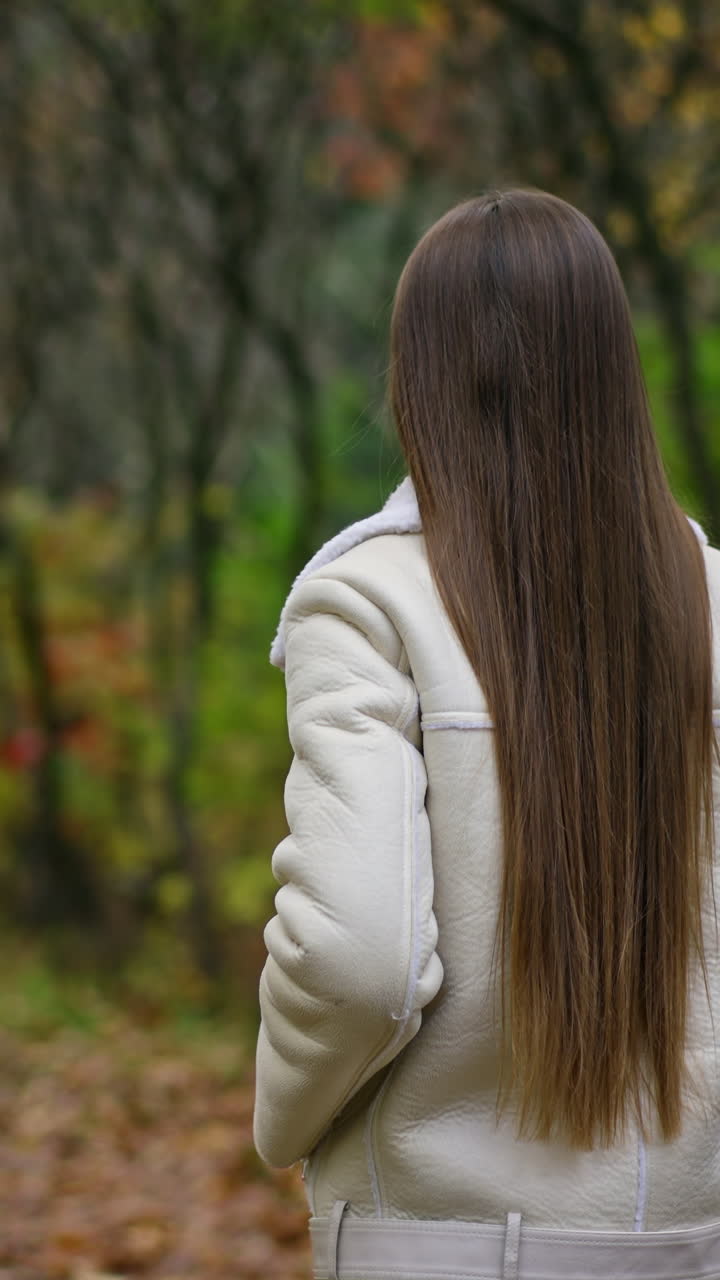 Following the lady with beautiful long dark hair walking in the park. Woman turns her head smiling to the camera. Autumn nature backdrop.
