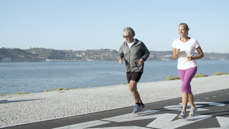 Sporty-people-jogging-on-asphalt-road-near-water-and-smiling