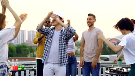 handsome young man in modern cap is dancing with friends at rooftop party, men and women are moving and clapping hands enjoying summertime and leisure.