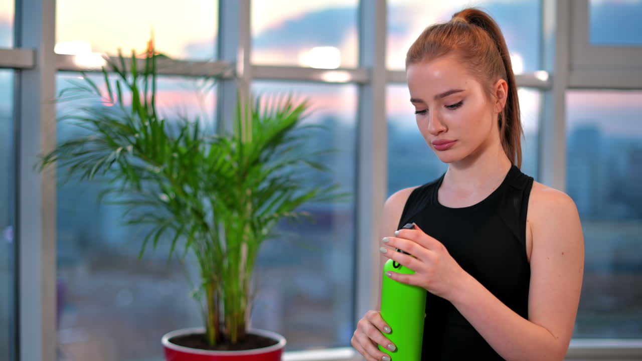 Young woman drinking water after workout with city on background. Indoor training in apartment, quarantine