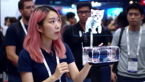 a presenter showcases innovative holographic technology with a 3d cat representation. attendees observe closely at a vibrant tech trade show filled with excitement.