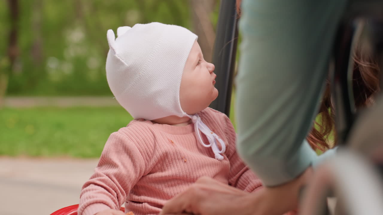 Baby Observing Caregivers By Wheelchair, Little Face Turned Toward Adults, Soft Hat And Pink Outfit, Attentive Gaze Captures Quiet Interaction, Park Path And Supportive Family Presence