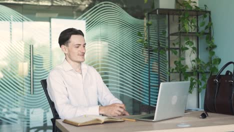 stylish man in business attire sitting at a desk with a laptop in a modern office, talking on a video call, giving a thumbs-up, making a heart gesture, and blowing a kiss