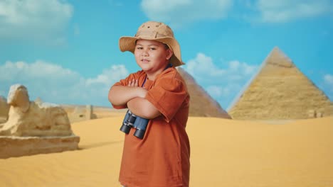 asian boy with a hat and binoculars crossing arms and smiling to camera while traveling in giza pyramid. boy researcher examines something, travel tourism adventure, side view