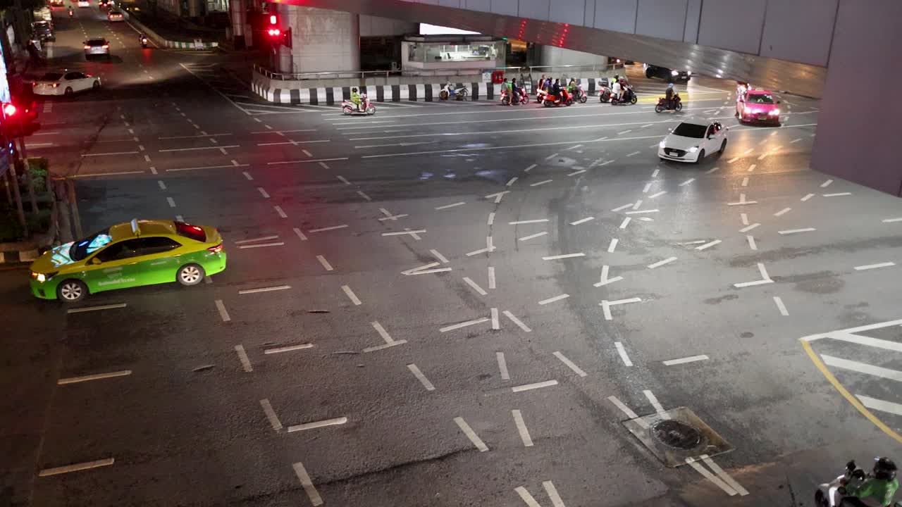 Cars and taxis navigate a well-lit Bangkok intersection at night, viewed from elevated perspective