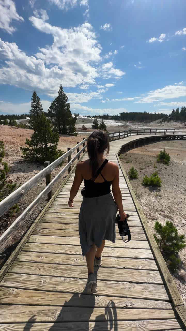 vista vertical de una fotógrafa caminando con una cámara por un sendero de madera en el parque nacional de yellowstone, estados unidos