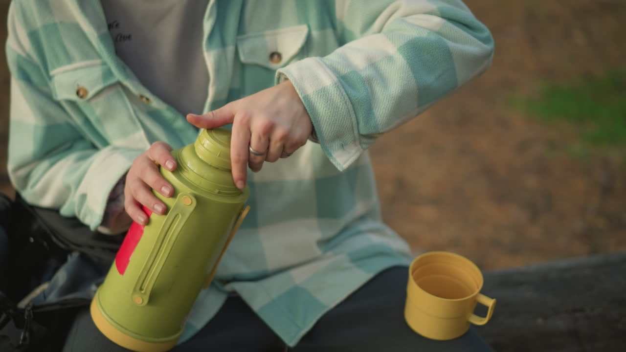 Male traveler unscrews thermos lid and prepares cup in closeup, focused hands working on yellow flask, tactile motion and threaded cap detail captured in natural outdoor light at a quiet forest rest