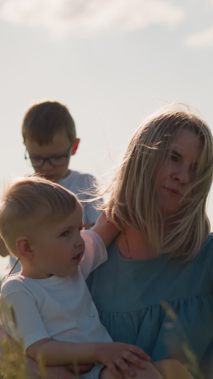 Close-up of a woman in a blue dress sitting on grass, holding a smartphone while filming herself. Her younger son sits on her lap, and her older son is focused on his phone behind them