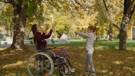 girl throws autumn leaves up above mother in wheelchair