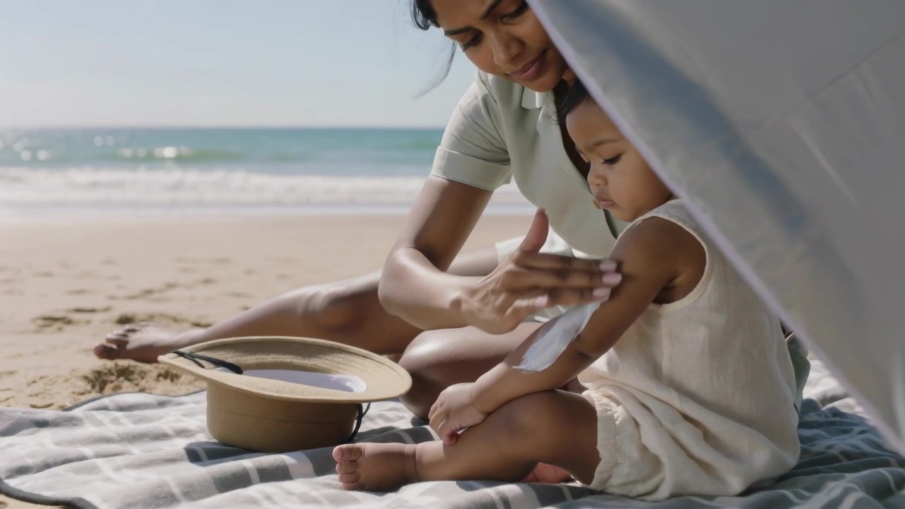 Touching arm, mother in green applying lotion rubbing toddler in romper on blanket for sun care