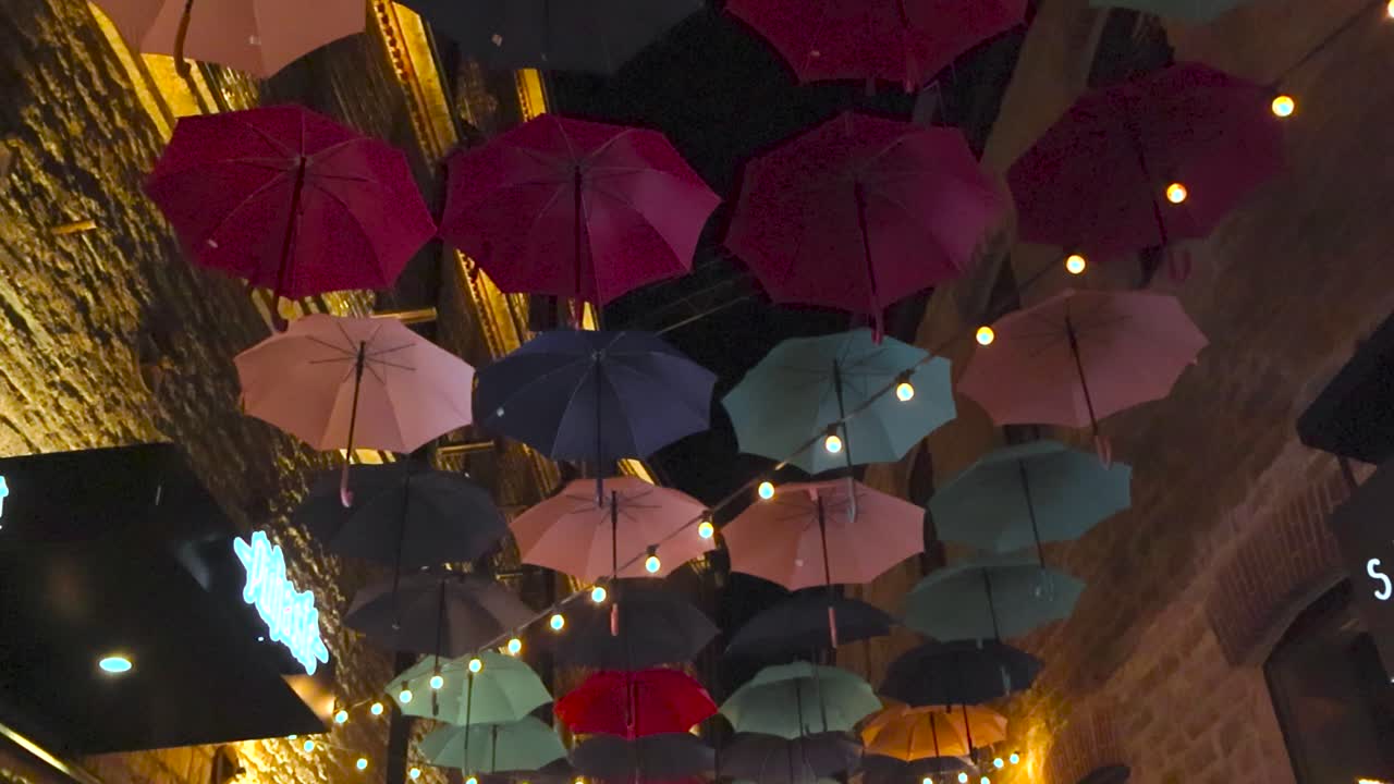 Gorgeous colorful umbrellas hanging over a narrow street in Rotermanni street or block in Tallinn during night or evening time. Restaurant are at the street where people are sitting drinking, eating.
