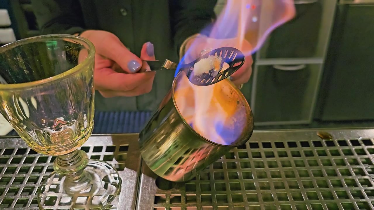 Primo piano di mixology notturna con fiamma blu, mani del barista, zolletta di zucchero, cucchiaio forato, shaker in metallo, calice vuoto e bancone del bar lucido in un cocktail lounge buio.