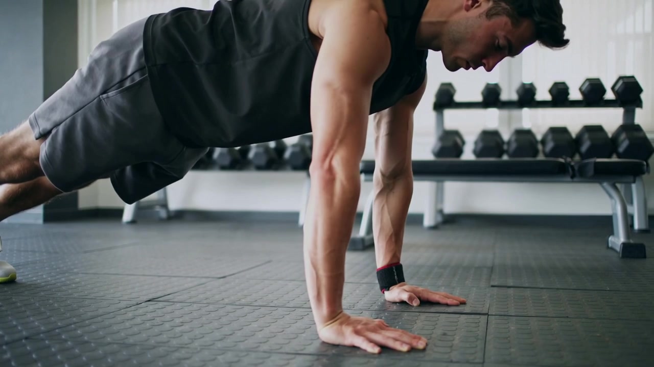 Uomo che inizia un plank alto, facendo flessioni sul pavimento della palestra per allenamento, indossando una canotta, con una rastrelliera per manubri.