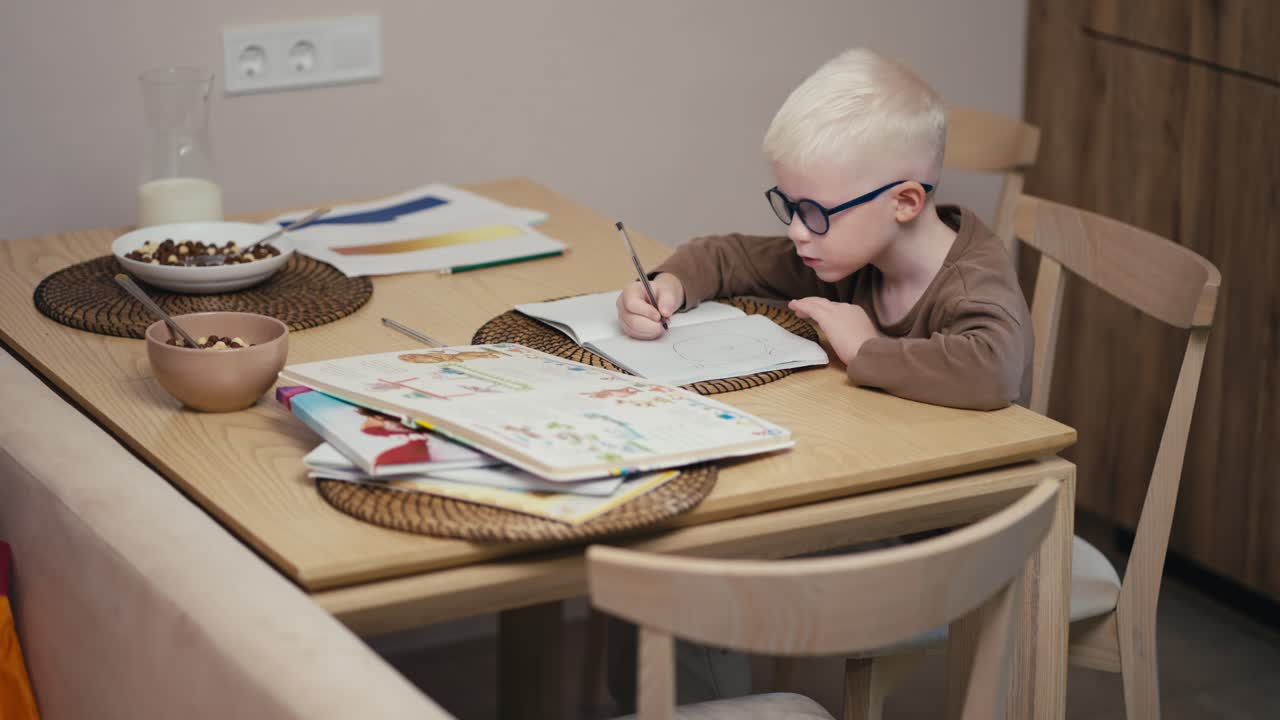 A little albino boy with white hair wearing blue glasses writes with a pen on his notebook and prepares his homework before the next day at school in a modern apartment in the kitchen