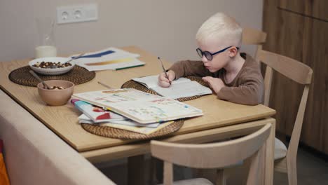 Ein-Kleiner-Albino-Junge-Mit-Weißem-Haar-Und-Blauer-Brille-Schreibt-Mit-Einem-Stift-In-Sein-Notizbuch-Und-Bereitet-Seine-Hausaufgaben-Vor-Dem-Nächsten-Schultag-In-Einer-Modernen-Wohnung-In-Der-Küche-Vor