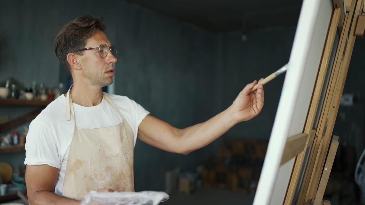 안경을 입은 젊은 행복한 예술가는 캔버스에 그림을 그리고 있습니다. 창의적인 사람의 일상 생활. 창의성 개념