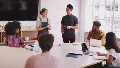 two millennial colleagues stand presenting to creative team in a meeting room