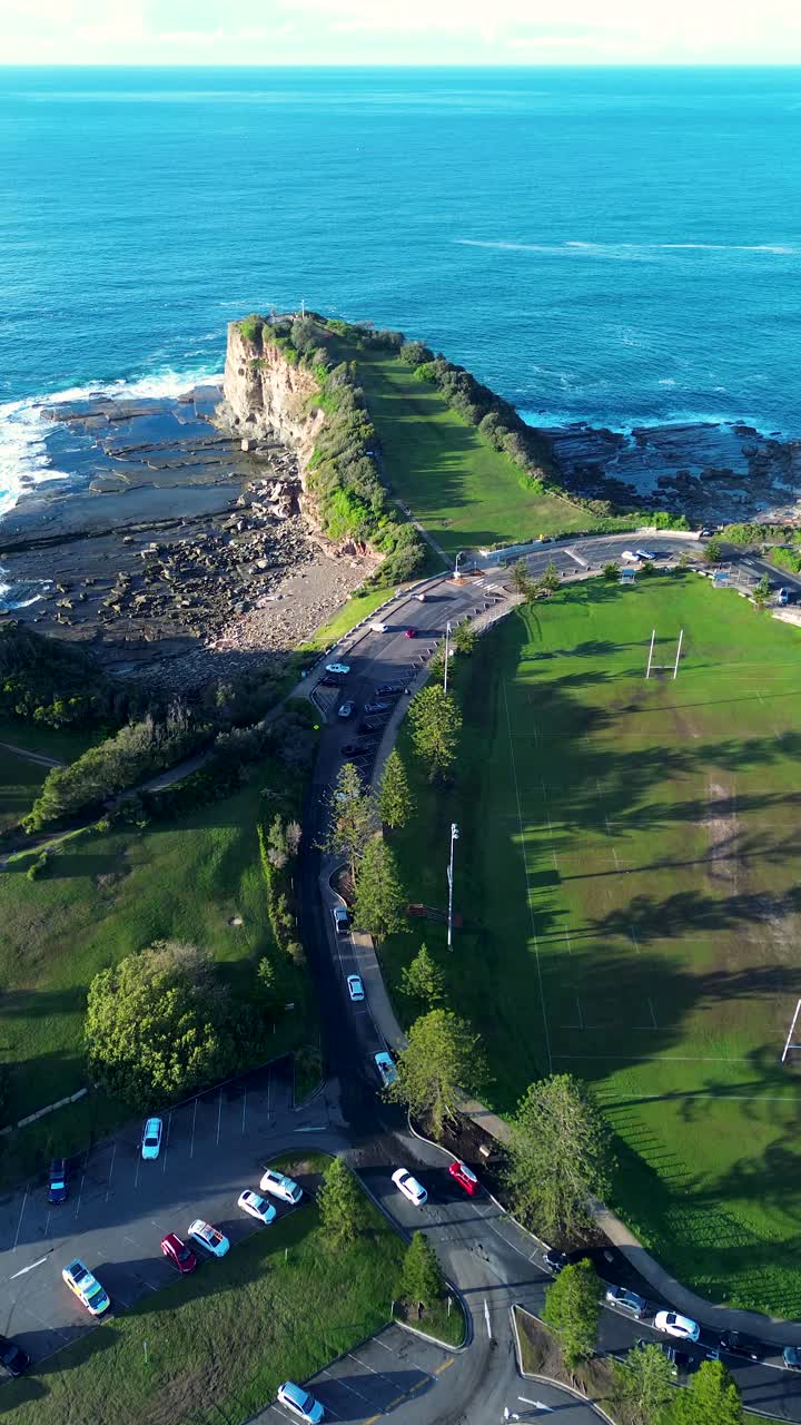 Aerial drone portrait of The Skillion cliffside headland Terrigal rocky hill landmark attraction near sports field oval and vehicles on road Central Coast Australia ocean bay travel tourism beachside