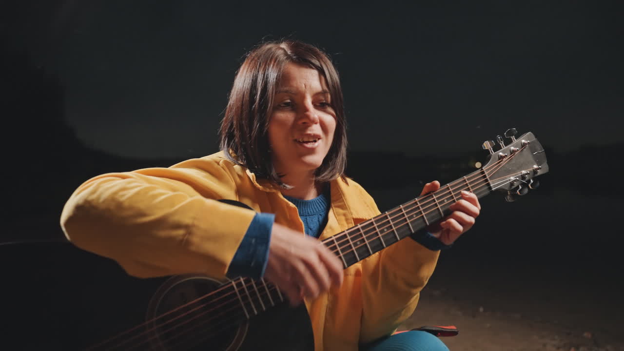 Giovane donna che strimpella la chitarra durante una notte in campeggio sulla spiaggia sabbiosa, primo piano di sorriso e risate, giacca gialla brillante, coro vivace e luce calda del fuoco, atmosfera acustica amichevole e performance espressiva.