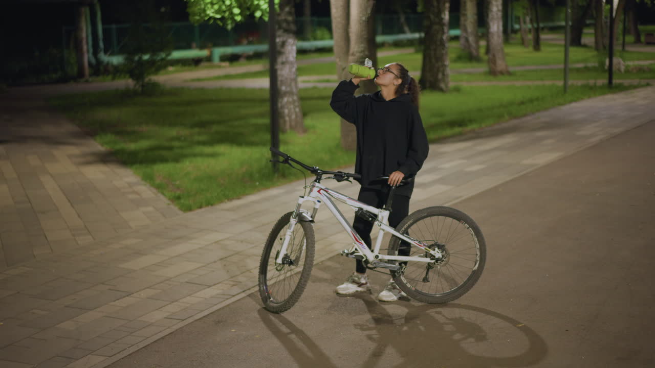 Woman Examines Phone Near Bicycle During Night, Individual Pauses To Review Route While Biking At Night, Female User Consults Mobile Device Beside Bicycle On Park Trail During Evening Hours