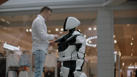 Un-Hombre-Con-Camisa-Se-Comunica-Con-Un-Robot-Blanco-Haciendo-Preguntas-Y-Presionando-La-Pantalla-Con-Los-Dedos