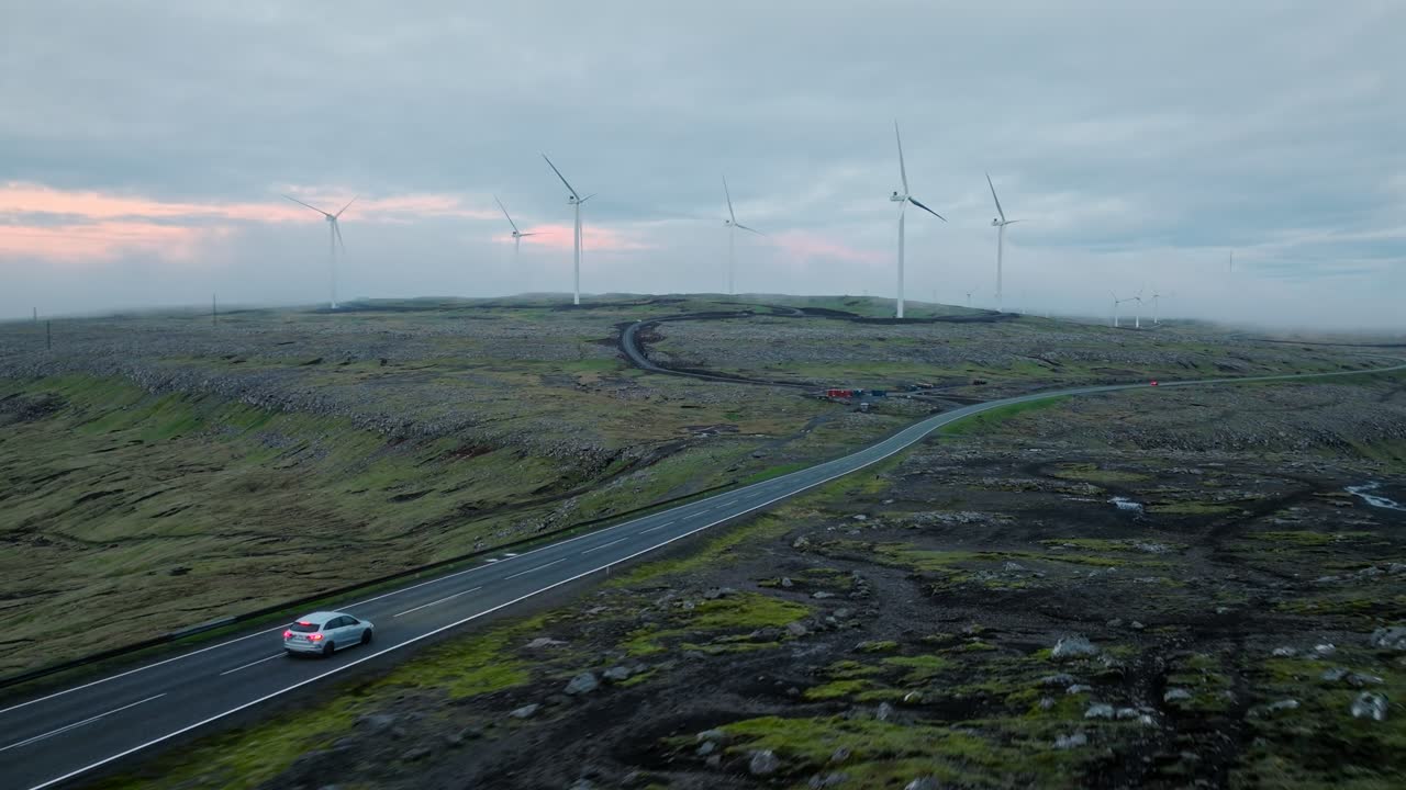 A car drives through a remote Faroe Islands landscape with wind turbines on a cloudy day