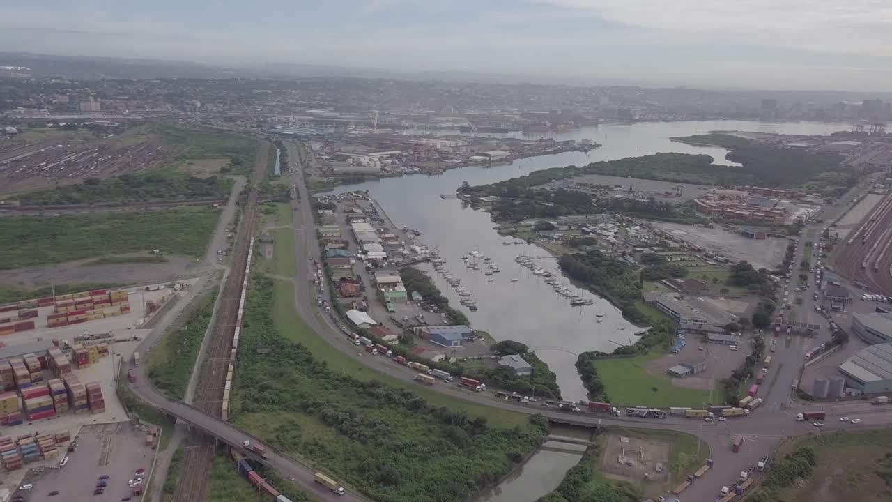 Aerial footage of Durban harbour with yachts and traffic consisting of trucks and moving trains over a highway with cargo container depot in view.