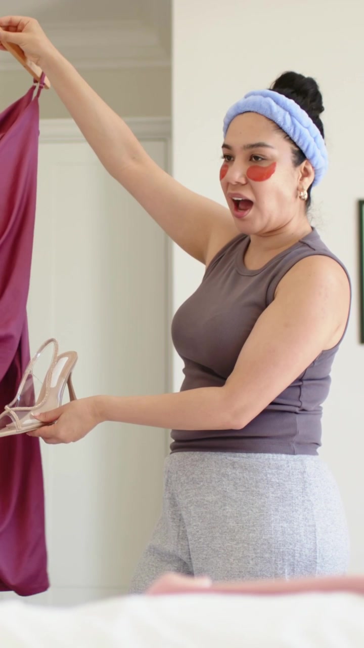 Vertical video: Stepping in woman in gray tank choosing outfit in bedroom holding dress and sandals